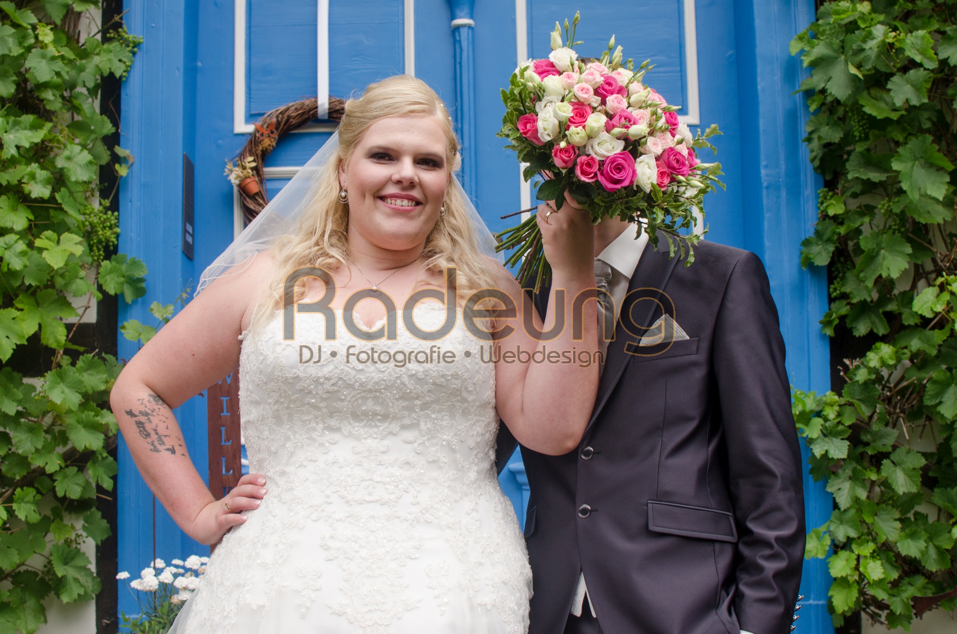 Ein Brautpaar steht vor einer blauen Haustür. Die Braut hält einen Blumenstrauß vor das Gesicht des Bräutigam.